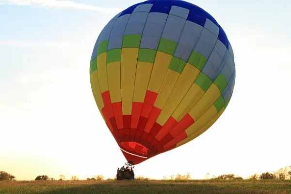 Personas subiendo a una canasta de un Globo aerostatico en tonos azules, amarillos y rojos, parado sobre el cesped. Esperando para comenzar su viaje en globo teotihuacan.