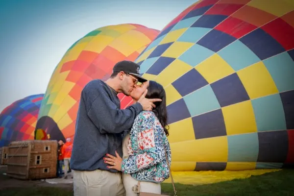 Pareja disfrutando del amanecer durante sus vacaciones en globo con vistas a paisajes naturales y globos aerostáticos elevándose en el cielo.