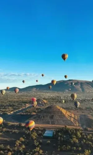 Turistas disfrutando el festival de globos aerostáticos mientras sobrevuelan las impresionantes pirámides de Teotihuacán iluminadas por el sol.